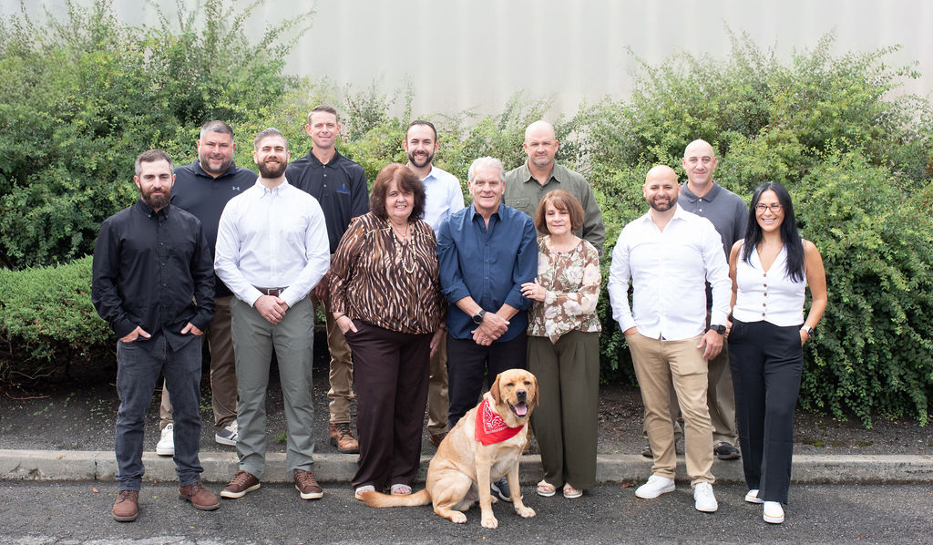 The twelve people of Montana Contracting, and Franky, standing together in front of a hedge and a concrete wall